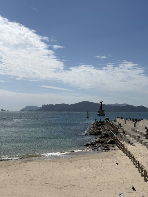 A scenic beach view featuring blue water, a sandy shore, and mountains in the distance, with a statue on a rock outcropping and people walking along a promenade.