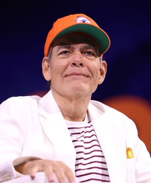 The image features a man wearing an orange cap sitting down while looking off-camera with his left hand resting on his lap. The background of the photo shows blue and orange tones creating a visually appealing contrast. In the surrounding area, there's an event where another person is talking or gesturing in front of the main subject. The main subject also has something written above their head that reads "MAX KEISER: “I'm raising my target price for Bitcoin from $220,000 to $2,200,000".