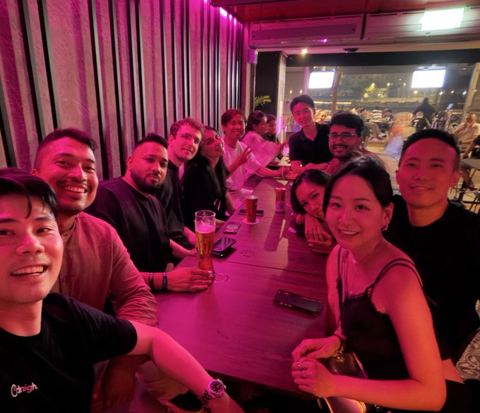 A group selfie of approximately ten people smiling at a restaurant table, bathed in pink/magenta lighting with beer glasses visible.