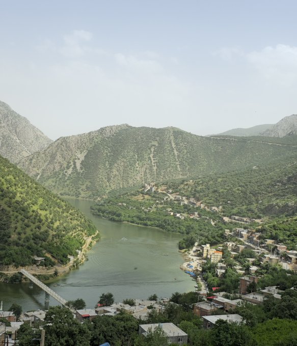 An aerial landscape view shows a green river winding through a valley surrounded by mountains and dotted with small village houses and bridges.