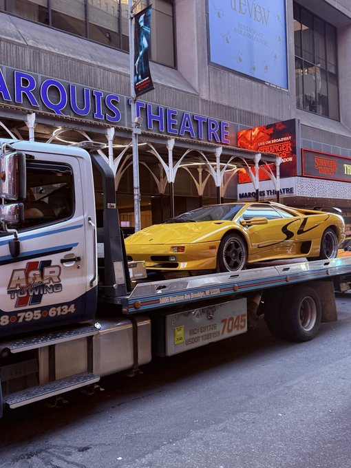 A yellow Lamborghini Diablo is loaded onto an 'AAR Towing' flatbed truck parked outside the Marquis Theatre, with a 'Stranger Things' marquee visible in the background.