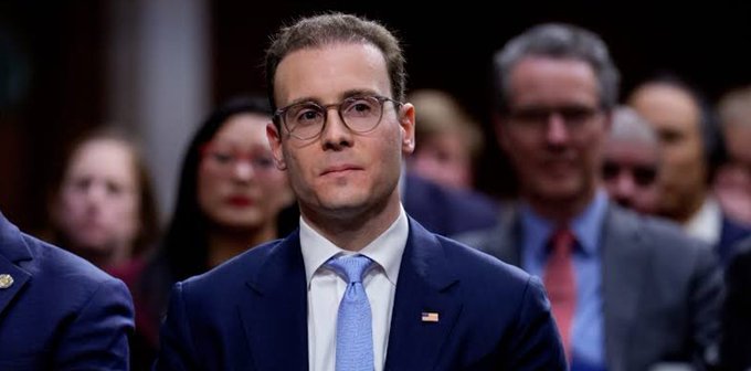 A man with short brown hair and round glasses, wearing a dark navy suit, light blue tie, and an American flag lapel pin, stands in a formal setting with a blurred audience behind him. The image appears to connect to the post by depicting a regulator or official, likely representing the CFTC's new leadership, whose serious expression reflects the agency's push for stricter crypto oversight and fraud enforcement.