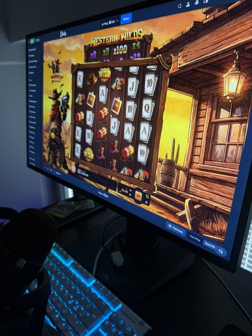 A photograph of a computer monitor displaying a Western-themed online slot game called "Western Wilds," with a microphone and illuminated keyboard visible in the foreground.