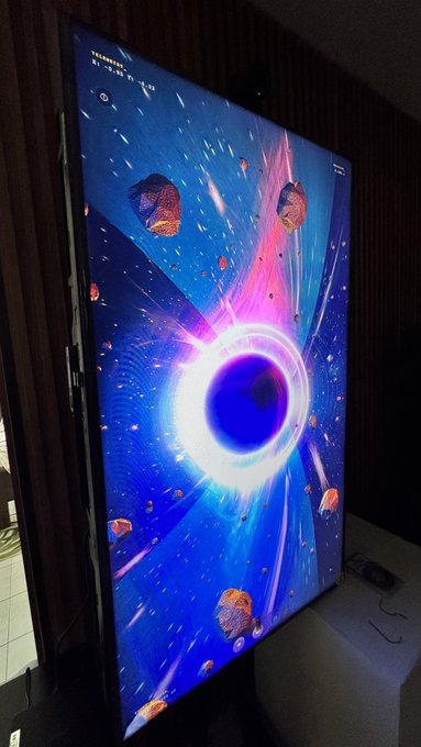 A large digital display shows a vibrant, interactive artwork depicting a cosmic scene with a central black hole surrounded by asteroids and pink-hued energy beams against a deep blue starfield. Visible text on the screen reads "FEBRUARY" along with a timestamp of "17:59:52," complementing the post about the GAIA BH3 supermassive black hole piece on @objktcom.