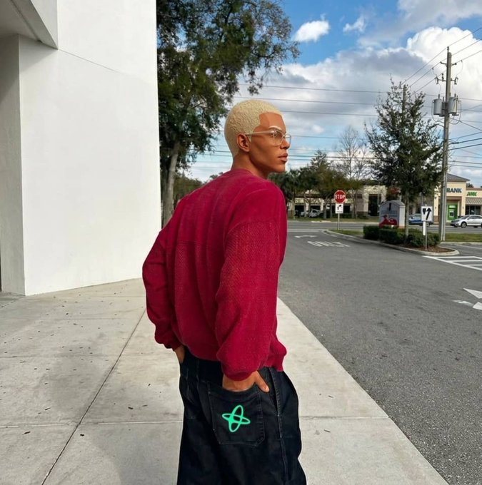 A person with short blonde hair wearing a red sweater and black jeans, standing on a sidewalk with a street intersection in the background.