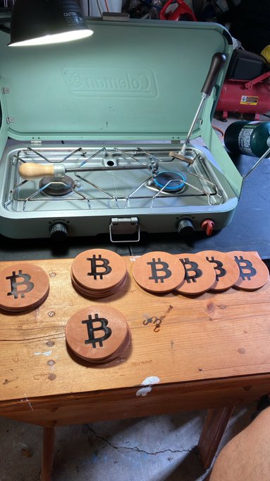 A green Coleman camping stove sits open on a workbench, with several wooden coasters engraved with the Bitcoin '₿' symbol arranged in front of it.