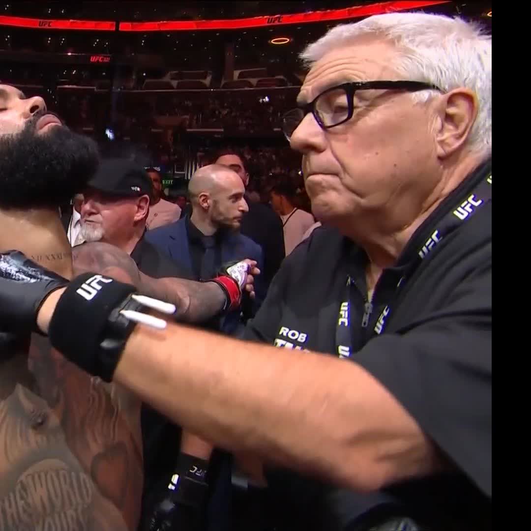 A referee wearing "UFC" and "ROB" branded gear prepares former champion Jon Jones, visible with his distinctive beard and tattoos, before his fight. The post's reference to "CryptoCom" likely refers to the Crypto.com Arena venue, and the image captures a behind-the-scenes moment of pre-fight preparation.
