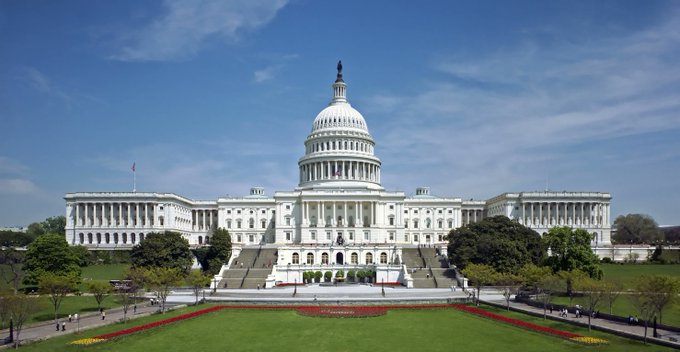 The image shows the United States Capitol building in Washington D.C., with a focus on its white facade, dome, and columns. The Capitol is surrounded by lush green trees, creating a serene atmosphere around the iconic landmark. In the background, there are people walking along the sidewalks near the Capitol grounds, adding to the bustling city vibe of the scene.