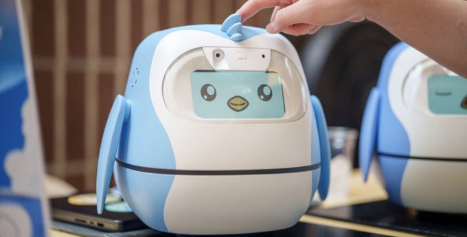 A close-up of a small blue-and-white penguin-shaped companion robot being touched on the head by a person's hand, with an identical unit visible in the background.