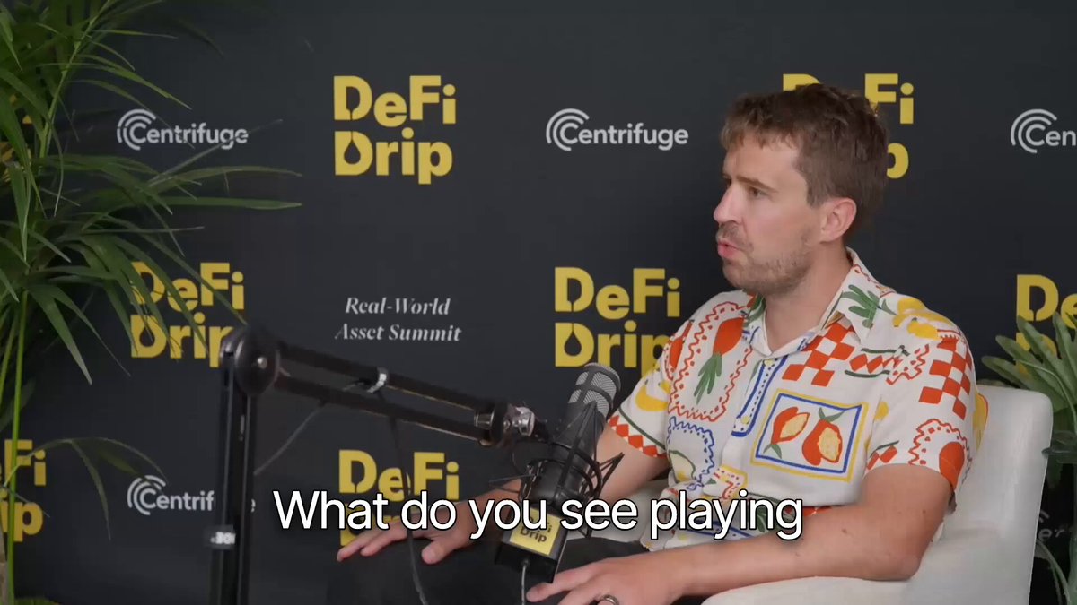 The image features a man sitting behind a microphone, likely participating in an interview or podcast discussion about Defi Drop. He is wearing a white shirt with colorful patterns. The backdrop of the scene includes a black wall adorned with yellow text that reads "Defi Drop" and "What do you see playing?". This suggests that the man might be discussing the concept of Defi Drop, which is a type of lending platform based on blockchain technology. The image also contains some text about DeFi Drip and how it has been featured in various media outlets such as @Morpho and @rwasummit.