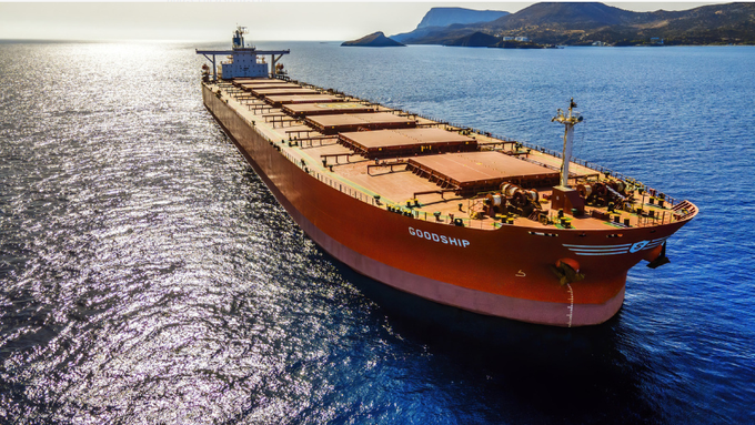 An aerial view of a large red cargo ship named "GOODSHIP" sailing on blue waters with mountains in the background.