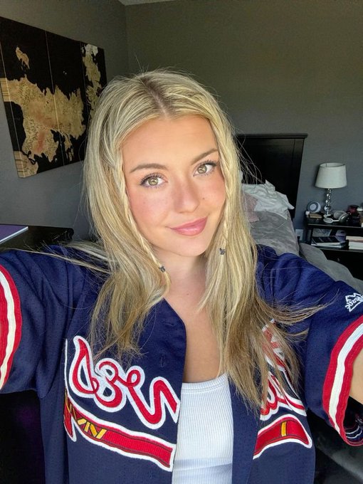 A young woman with long blonde hair taking a selfie, wearing a dark blue Atlanta Braves MLB jersey over a white top, in a bedroom with a map on the wall.