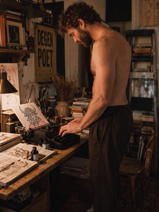 A shirtless bearded man types on a vintage typewriter in a dimly lit artist's studio, with a 'DEGEN POET' poster visible in the background alongside books and art supplies.