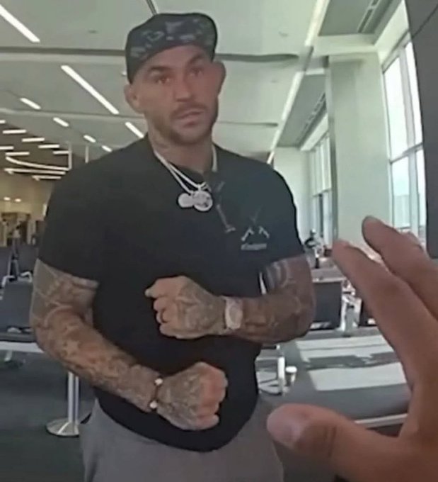 A muscular man with tattoos, wearing a cap, black t-shirt, and gold chains, flexes in a gym while another person's hand gestures toward him in the foreground.