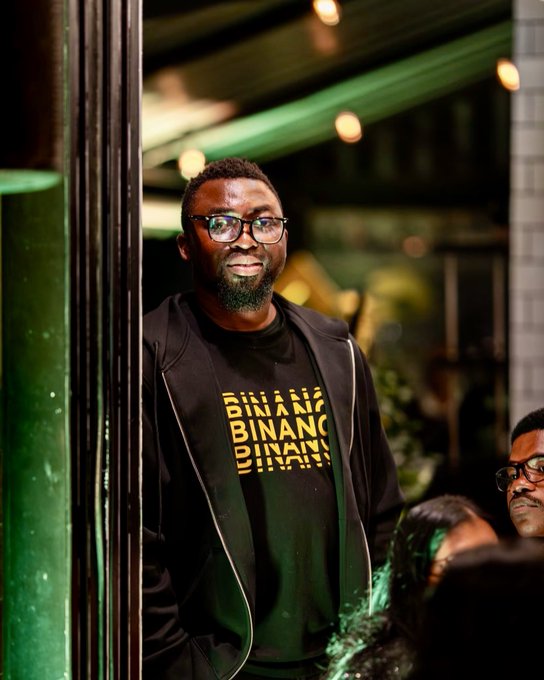 A photograph of a smiling bearded man wearing glasses and a black jacket over a t-shirt that reads "BINANCE" repeated in yellow text.