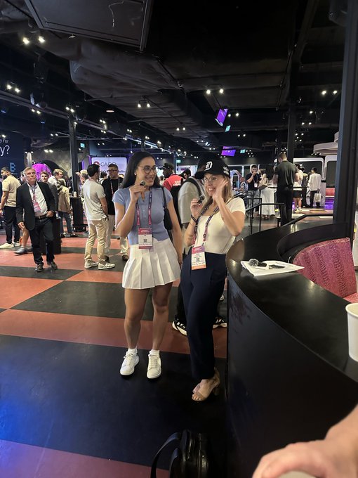 Two women pose for a photo at what appears to be a busy finance conference, both wearing lanyards and holding microphones, with the woman on the right wearing a "D" cap in reference to the Doginal Dogs community mentioned in the post. The venue features a modern lounge area with a checkered floor, low lighting, and a crowd of attendees networking in the background.