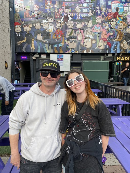 A smiling couple poses together at an outdoor venue featuring a large colorful mural depicting cartoon monkey characters on the building behind them.