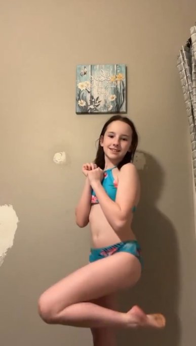The image shows a young person posing playfully with arms crossed and one leg lifted in a room with a floral artwork on the wall behind them. They are wearing a two-piece swimsuit in teal, pink, and yellow colors.