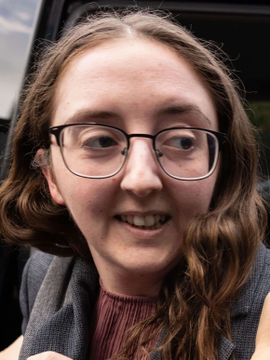 A smiling young woman with glasses and long brown hair is pictured in a close-up selfie, likely representing Caroline Ellison mentioned in the breaking news about FTX-related individuals being released from jail while facing new five-year trading bans from the CFTC.