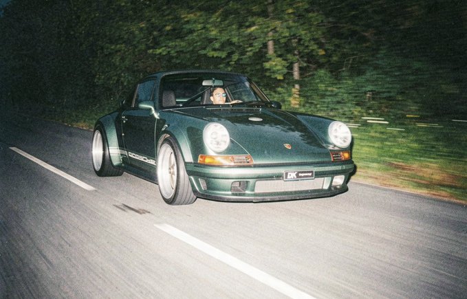 A dark green Porsche 911 sports car driving on a forest road at night, showing motion blur and wearing an 'LK' license plate.