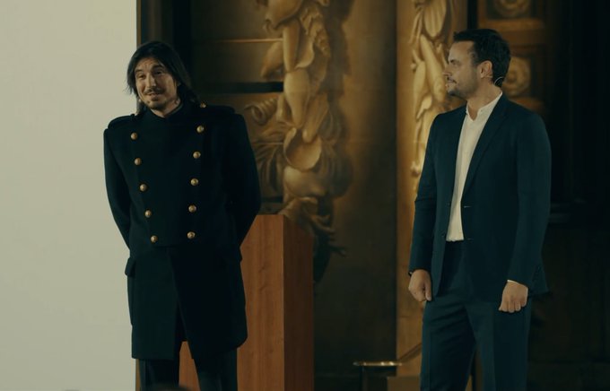 Two men in dark formal attire standing in an ornately decorated interior with golden sculptural reliefs on the walls.