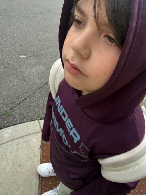 A young person wearing a maroon hoodie with "UNDER ARMOUR" text looks up at the camera from a low angle, standing on a sidewalk.