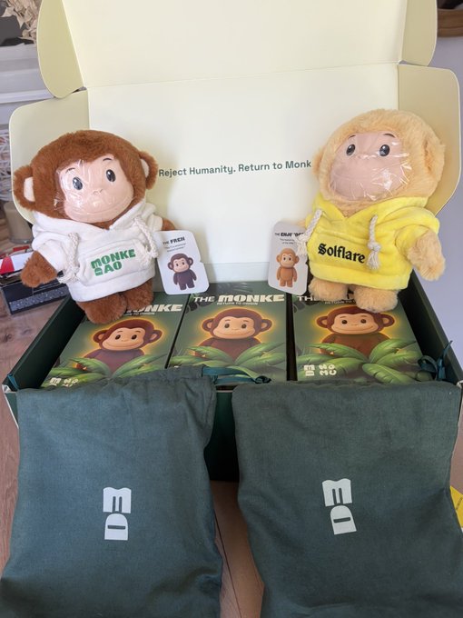 An open box containing 'Reject Humanity. Return to Monk' merchandise featuring two plush monk dolls (one in 'Bonke Dao' hoodie, one in 'Solflare' yellow), green 'DB' bags, and Solana (SOL) branded cards.