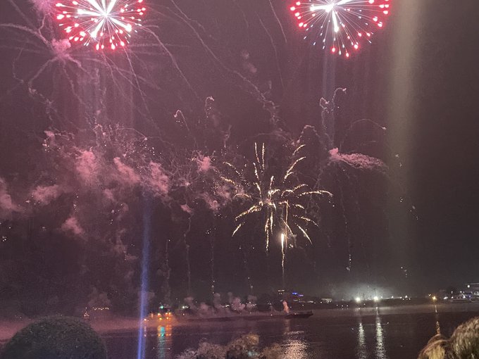Red and white fireworks burst over a body of water at night, with silhouetted spectators watching from the shoreline.