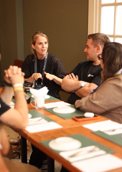 A woman with a lanyard speaking and gesturing at a restaurant table while a man in a black t-shirt listens with arms crossed, with other people seated around them.