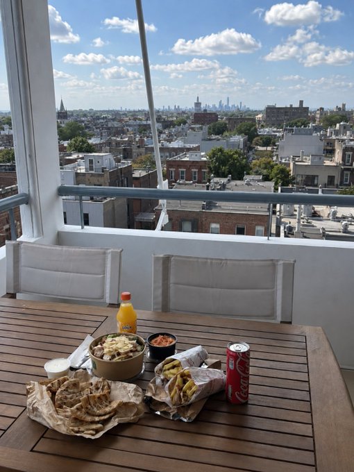 A casual weekend meal of wraps and fries with Coca-Cola sits on a rooftop table overlooking a city skyline, providing a perfect chill backdrop for the poster's relaxed "gmeow all" vibes—tucking into food before heading out to "buy some coins." The Coca-Cola can is clearly visible on the table.