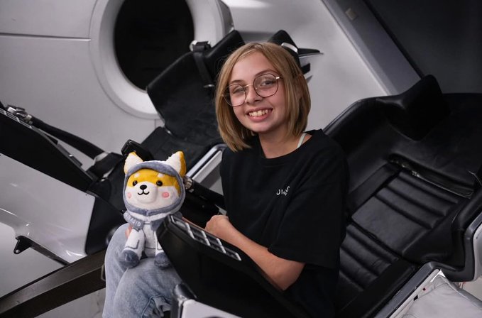 The image features a young girl sitting in a chair wearing glasses and holding a stuffed animal. She has short blonde hair and is smiling at the camera, giving off a happy vibe. The background of the photo shows a white car with a black interior, which appears to be a part of an advertisement or promotional material for Asteroid.