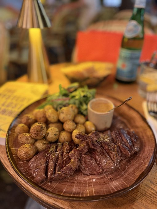 A hearty meal of sliced steak and roasted baby potatoes served on a wooden platter with greens and sauce, with a green glass bottle in the background.