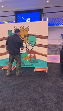 An artist wearing a dark hoodie and cargo pants stands with his back to the camera, painting a cartoon bear on a canvas at what appears to be a live event.