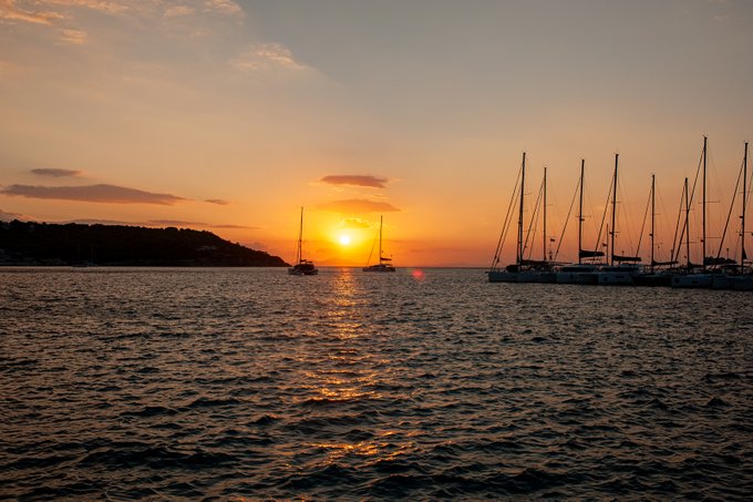 A scenic sunset photograph over the ocean with sailboats anchored on the right side and a hilly coastline visible on the left, the sun reflecting golden light across the water.