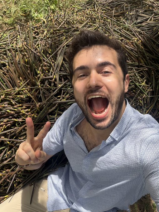 A man with a beard sits outdoors near dried vegetation, making an excited open-mouthed expression and holding up a peace sign.