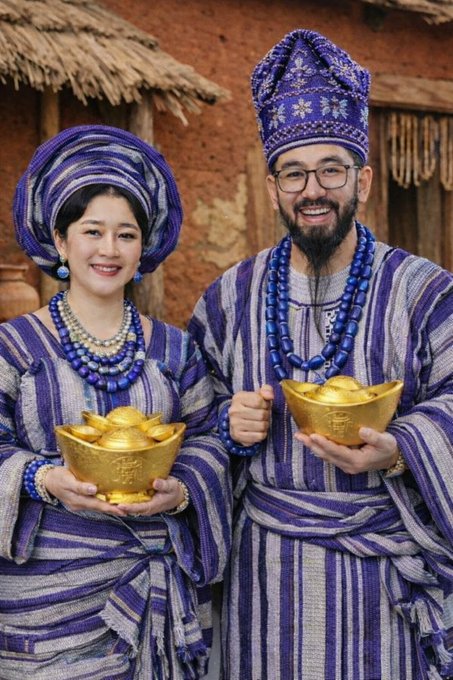 A smiling couple dressed in ornate blue and purple striped traditional attire with matching headwraps and beaded necklaces pose while holding gold ingot bowls (yuanbao) embossed with Chinese characters symbolizing wealth and prosperity, aligning with the Pi cryptocurrency post's theme of acquiring major brand goods through the Pi ecosystem.