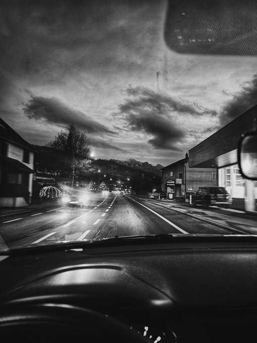 A black-and-white photograph taken from inside a car driving down a wet street at dusk, showing a dramatic cloudy sky, buildings, and the dashboard with a side mirror visible.