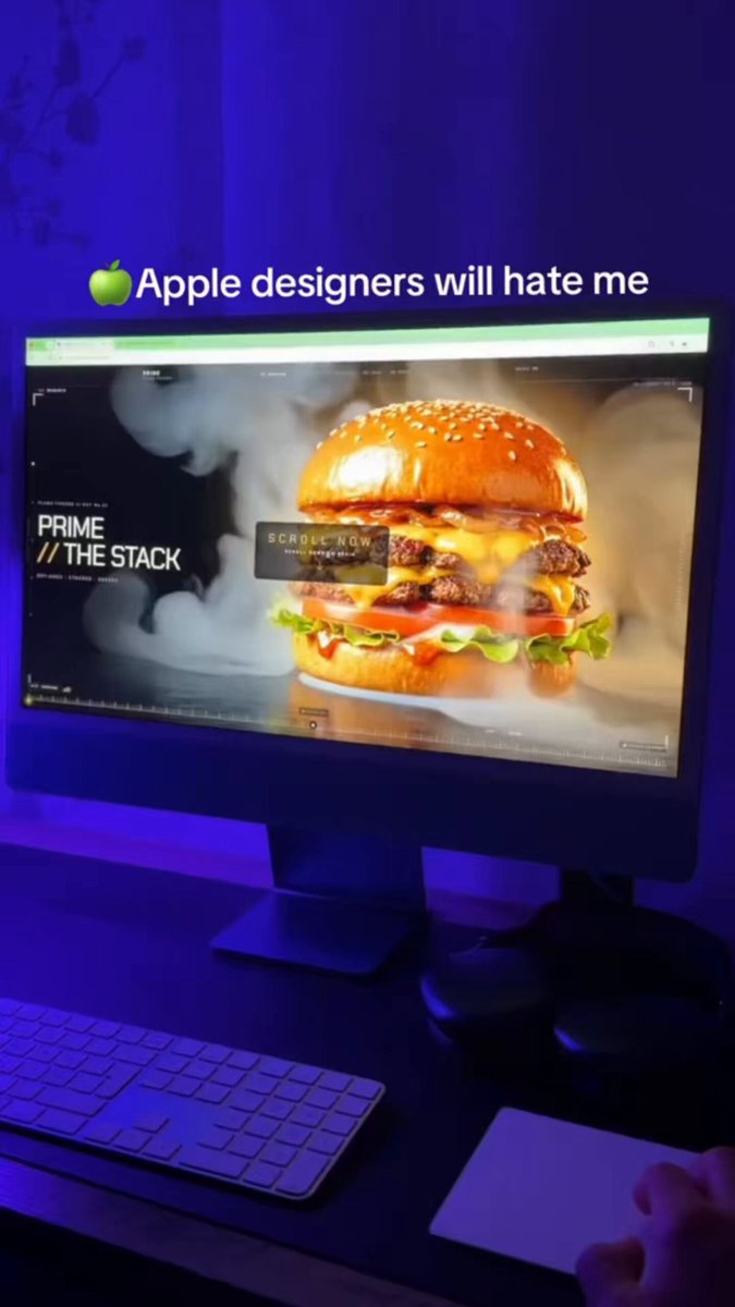 The image shows a computer monitor displaying an advertisement for Apple designers who will hate me. The screen features a large hamburger with cheese and lettuce as its toppings. A keyboard and mouse are placed next to the monitor, suggesting that this scene takes place in a workspace or home office setting.