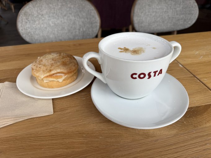 A Costa coffee cup with a cappuccino sits on a table next to a small pastry on a white plate.