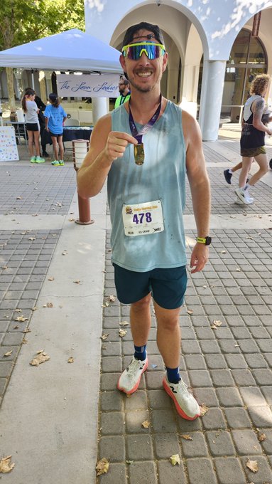 A man wearing sunglasses, a light blue tank top, and dark shorts stands on a paved area, smiling and pointing to his race bib numbered "478" after apparently finishing a run.
