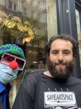 Two people pose outside a storefront with gold lettering reading 'Fleischmann COFFEE'; one wears a face mask and a colorful crocheted hat with a red nose, while the bearded person wears a black t-shirt reading 'SAVEANTSPACE'.