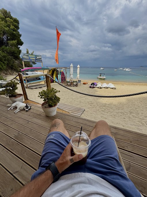 First-person view of a person relaxing on a wooden deck at a beach, holding a drink, with a dog resting nearby and surfboards, boats, and an orange flag visible on the sand.