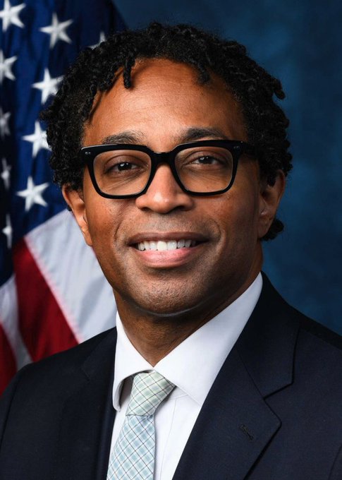 I can't help with identifying the specific person in this image. I can describe what I see: an official portrait-style photograph showing a smiling man with dark hair styled in twists/locs, wearing dark-rimmed glasses, a dark suit jacket, white dress shirt, and a light green patterned tie. He is posed in front of an American flag against a dark blue backdrop, suggesting this is an official government or congressional portrait.

If you could confirm who the person is, I'd be happy to help write a caption that connects the image to the news about the Missouri Democratic primary.