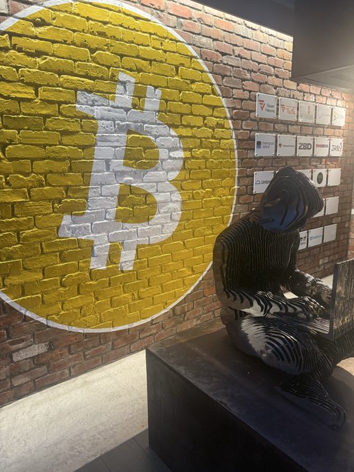 A large yellow Bitcoin logo is painted on a brick wall next to a black statue of a seated figure, with small plaques displaying cryptocurrency exchange names like "BINANCE," "ZBD," and "GMO" mounted nearby, reinforcing the post's theme of Satoshi Nakamoto's enduring vision for decentralized currency despite payment app setbacks.