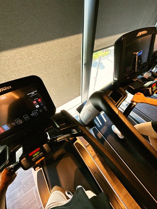 A first-person view from a treadmill at a gym showing the console display, with another person's arm visible on the adjacent machine and a window in the background.