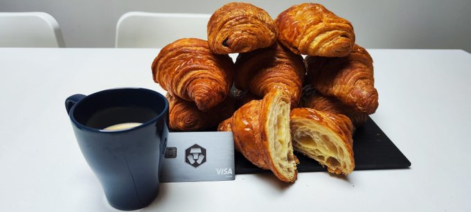 A breakfast scene shows a stack of croissants on a dark slate board next to a black Visa card and a blue mug of coffee.