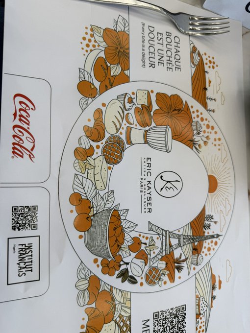 The image shows a placemat or menu from a French restaurant featuring "CHAQUE BOUCHÉE EST UNE DOUCEUR" (Every bite is a delight) and the chef's name "ERIC KAYSER," illustrated with autumn-colored foods, flowers, and the Eiffel Tower, along with a Coca-Cola logo and "FNTSUT FRANCAIS" branding. The "Touch some grass" joke plays on the rustic French countryside imagery of grass, flowers, and pastoral scenes depicted in the decorative illustration surrounding the central plate design.