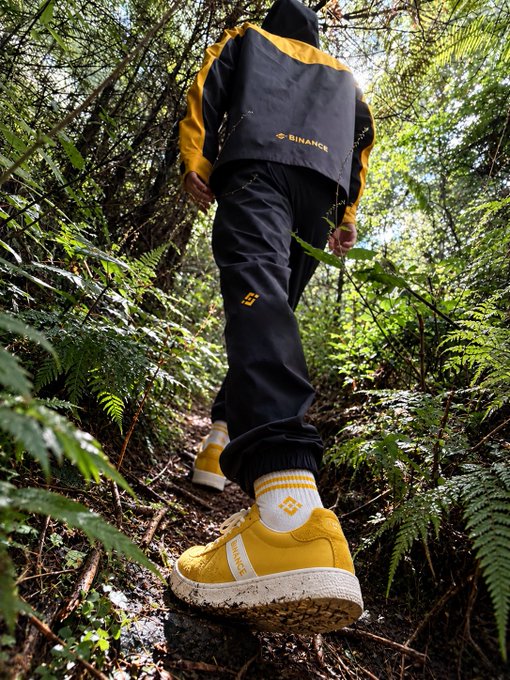 A hiker walks through a lush forest wearing Binance-branded black and yellow apparel, including a tracksuit with the "BINANCE" logo visible on the back and matching yellow sneakers, promoting the brand's new Mid-Autumn Festival merchandise.