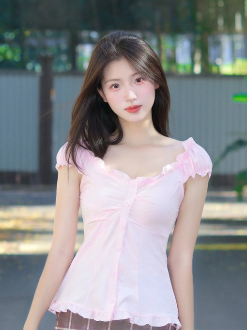 A young woman with long dark hair wearing a pink ruched top stands outdoors with greenery blurred in the background.