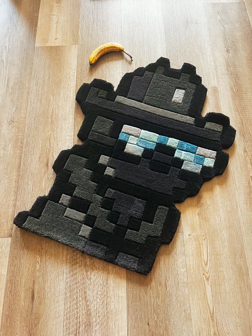A pixelated tufted rug shaped like the iconic Doginal Dogs pixel art dog lies on a wooden floor next to a banana for scale. The rug visually represents the Doginal Dogs meme, with the banana referencing the well-known "This is fine" / banana-for-scale meme aesthetic commonly associated with the Doginal Dogs community.